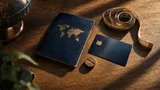 Overhead shot on a plain oak desk: a leather passport, a matte credit card, and a small brass turnstile token arranged beside a coiled measuring tape, lit by soft window light with long shadows suggesting scale and quiet risk.