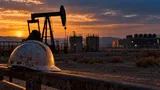 A pump jack silhouette stands against a hazy desert sky at dusk, its steel beam frozen mid-stroke beside a cluster of weathered storage tanks.