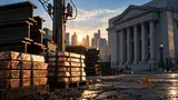 A sunlit industrial yard at dawn: stacked steel beams and raw copper ingots rest beside a weathered utility pole, its ceramic insulators catching the light, while in the soft-focus background a bank building's granite façade stands unlit an