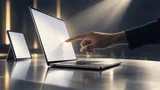 A silver laptop with a glowing blank screen and a fingertip hovering near its display, beside a smaller upright tablet, set against a dark softly lit studio backdrop suggesting an evening product launch event.