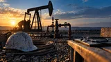 A sunlit industrial scene: a steel oil pump jack silhouetted against a dawn sky, its pipeline fittings glinting with dew, stands beside a weathered hard hat and a coil of thick cable resting on gravel.