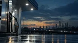 A dusk-lit petrol station fuel pump on a rain-slicked forecourt with a motorway and hazy city skyline in the background, evoking rising fuel costs and economic pressure.
