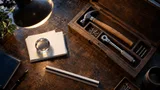 A top-down shot on a worn wooden desk: a wooden toolbox lies open, its compartments holding a hammer, a few loose screws, and a small torque wrench, each tool resting in its own molded slot.