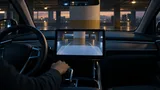 Interior view of an EV in reverse at dusk, showing a dashboard screen where a translucent overlay partially blocks the rearview camera image of a parking garage pillar, while the real pillar is visible through the back window above.