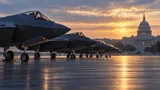 A row of F-35 fighter jets parked on a tarmac at dawn, with a government building silhouetted softly in the distant background.